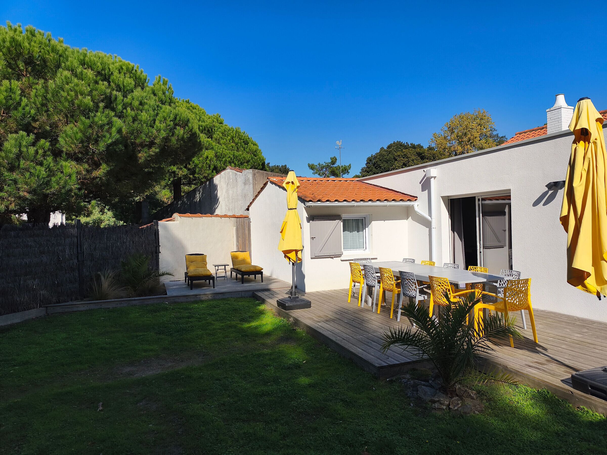 Une terrasse avec une table et des chaises, un parasol jaune et un petit coin jardin.