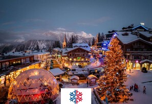 Ein mit Schnee bedecktes Dorf mit einer markanten Kuppelbauweise, einem großen, beleuchteten Weihnachtsbaum und einer Bergkulisse.