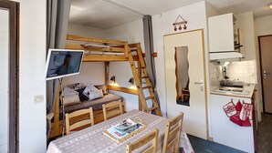 Quarto compacto com beliche, escrivaninha com monitor de computador, pequena cozinha e área de refeições com mesa e cadeiras.