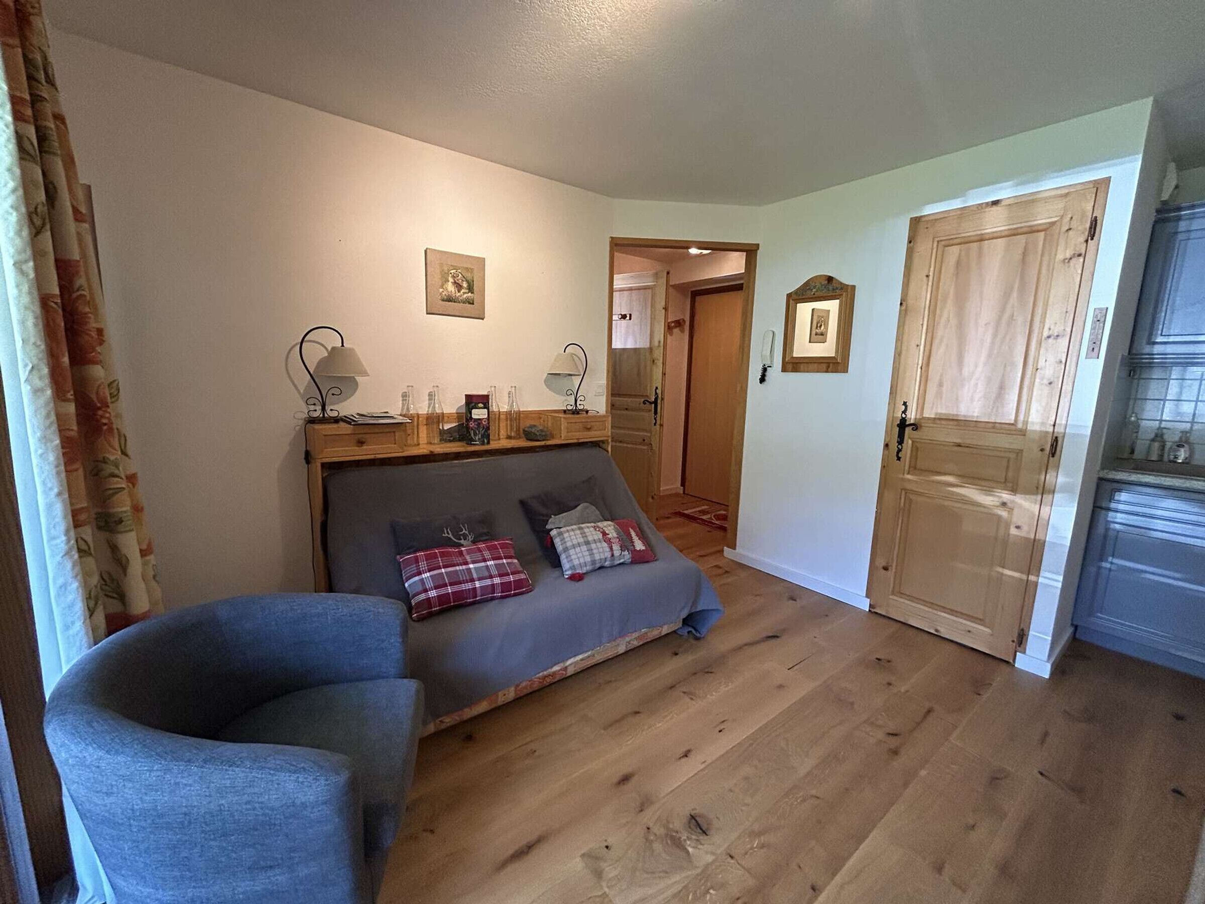 Una habitación acogedora con sofá, una silla, suelo de madera y puerta que da a una cocina.