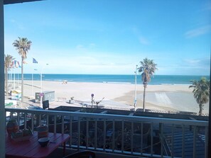 Blick auf einen Strand vom Balkon aus, mit Palmen, wolkenlosem Himmel und sandigem Ufer.