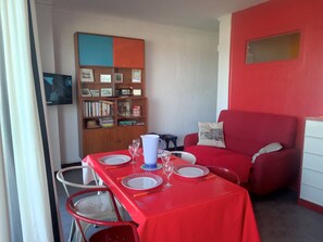 A dining area with a red table set for four, a bookshelf with various items, and a red sofa. - Spacious apartment sea view - 4 guests (Canet-Plage)