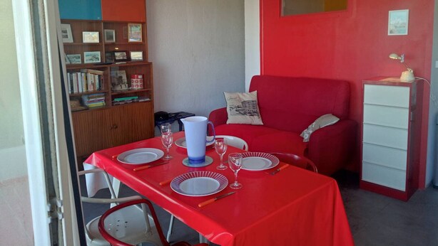 Un comedor con una mesa roja preparada para dos, una estantería, un sofá rojo y un mueble bajo con una lámpara.