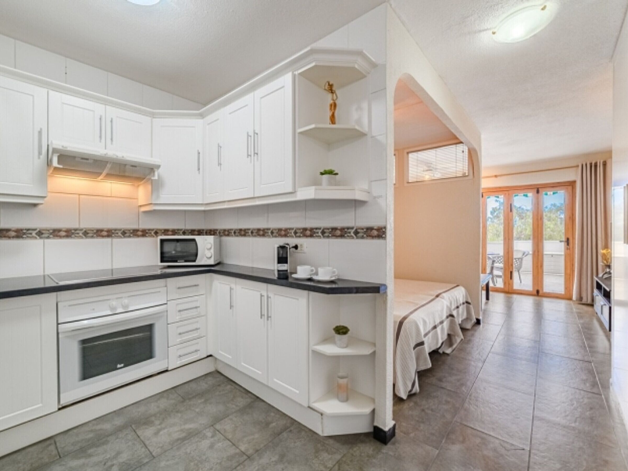 A modern kitchen with white cabinetry, a microwave, and a coffee machine. There is a small shelf with a plant and a candle. The kitchen leads to a dining area with a table and chairs.