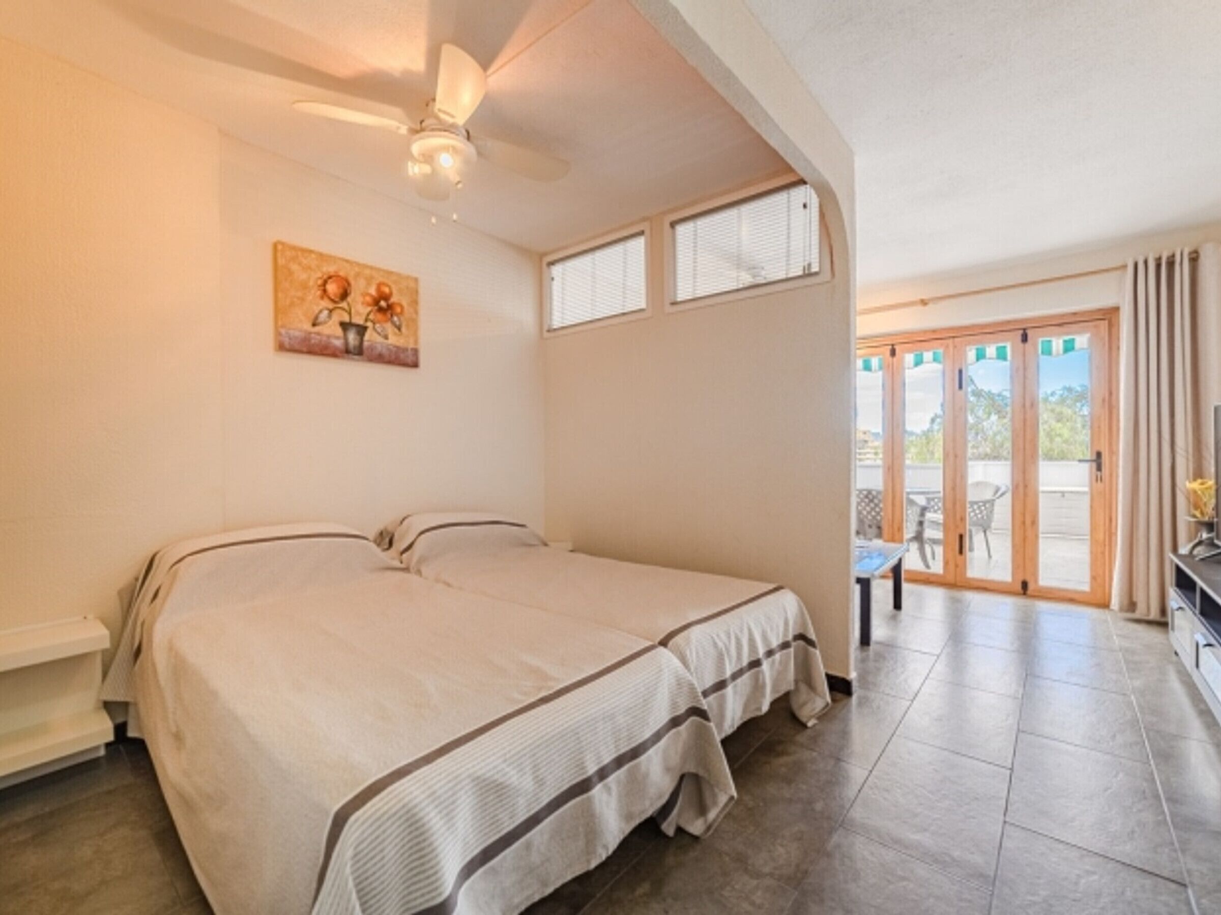 A bedroom with a bed, a ceiling fan, a painting, and a view through a glass door to an outdoor area.