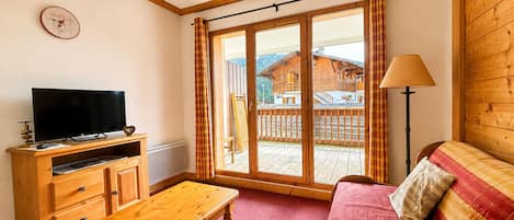 Ein Zimmer mit einer hölzernen Bank, einem Fernseher auf einem Holzgestell, einer Couch mit einem roten Kissen und Blick auf ein Holzgebäude durch eine Glastür.