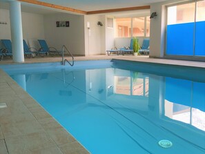 Una piscina interior con agua cristalina, rodeada de sillones de descanso y un suelo de baldosas.