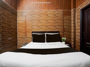 A hotel room with a wooden paneled wall, a bed with white bedding, a bedside table with a lamp, and a door. - Deluxe Hindon Airport Ghaziabad (Ghaziabad)