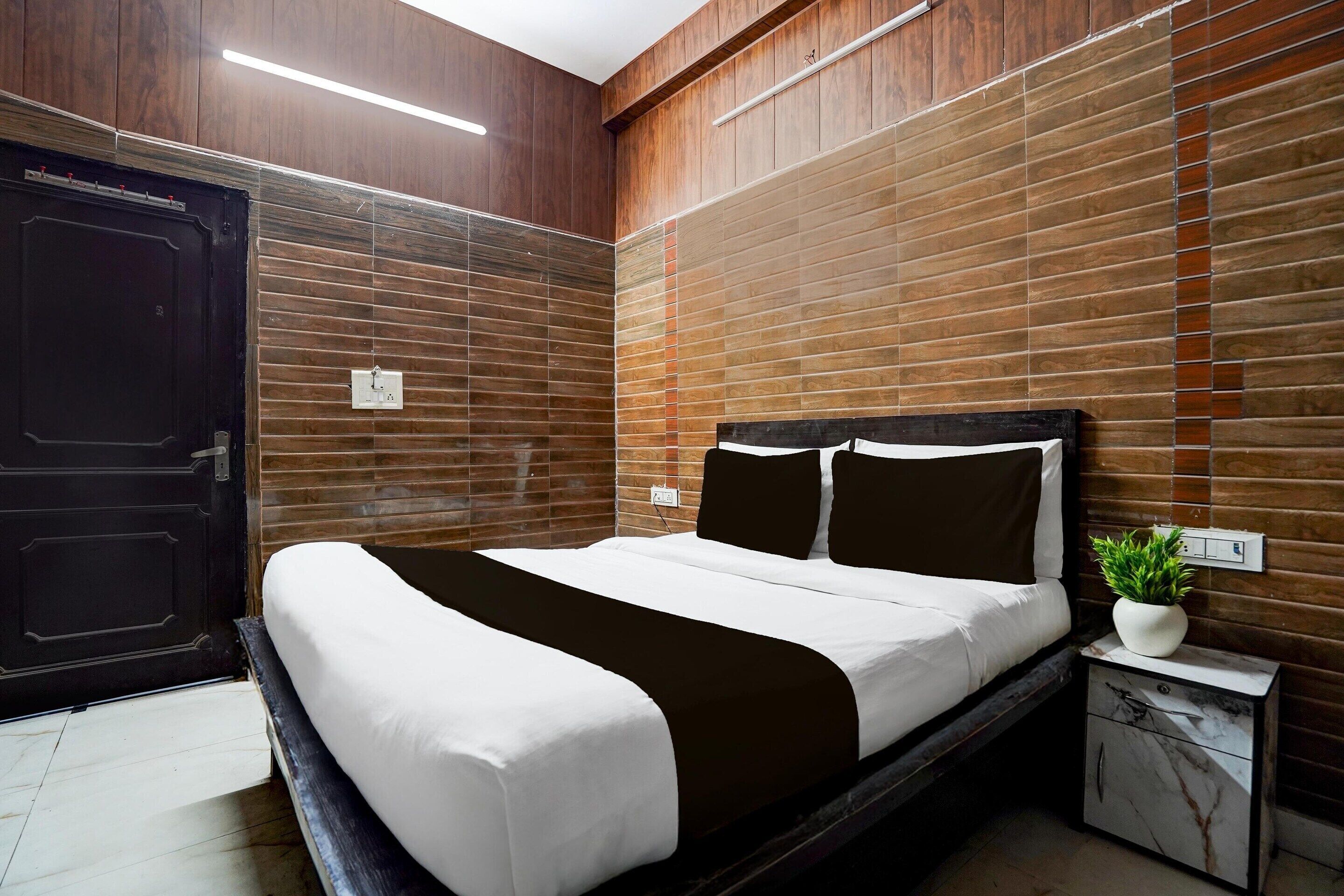 A modern hotel room with a large bed, a nightstand, a potted plant, and wooden paneling on the walls.