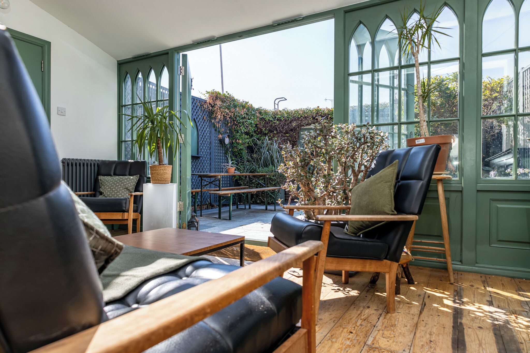 A sunlit room with a view of a garden, featuring a black leather sofa, a wooden table, and a chair.