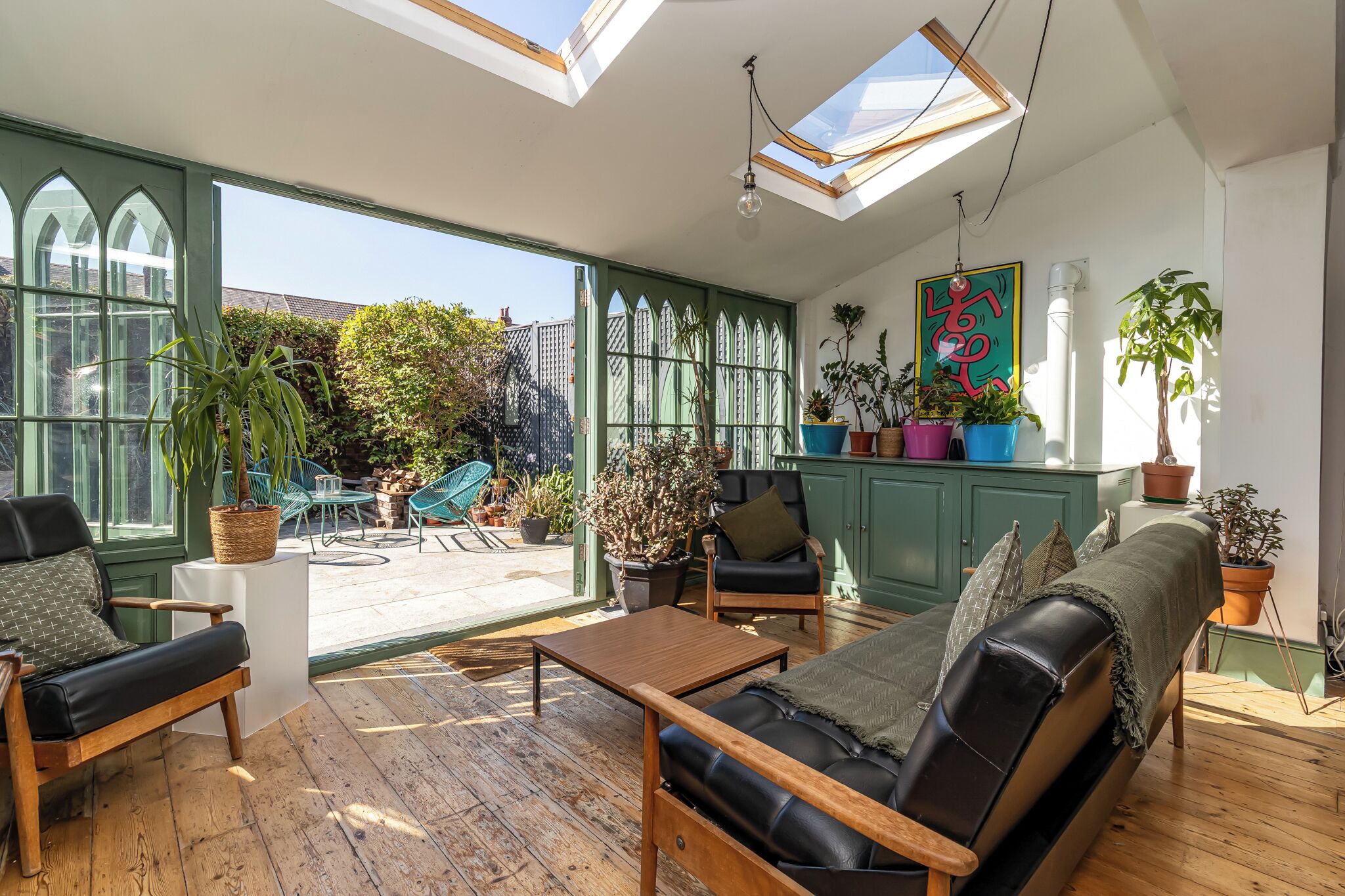 A sunlit room with a wooden floor, a sofa, two chairs, a table, and a cabinet with potted plants.