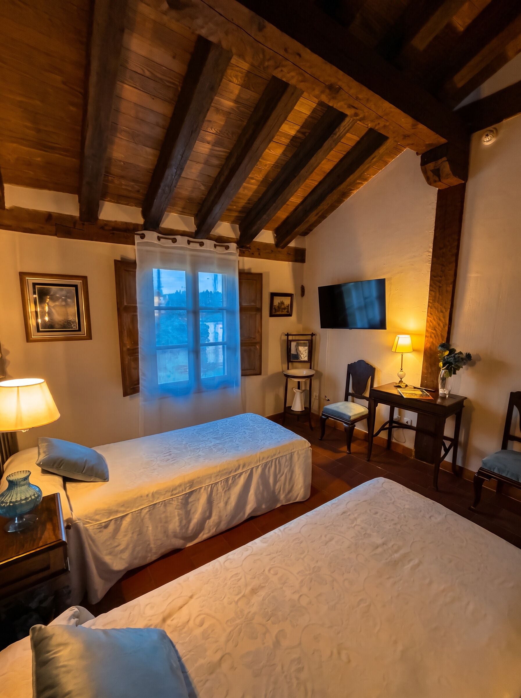 A cozy room with two beds, a desk, a chair, a TV, and a window with curtains.