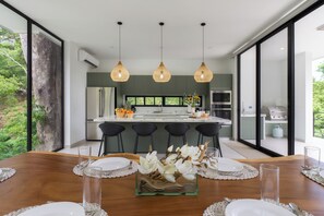 A modern kitchen with a central island, black bar stools, and a dining table set for six with white plates and clear glasses. - Casa Joanna | Luxury Jungle Retreat in Pacifico Resort | Private Pool (Pacifico Resort Lot 35)