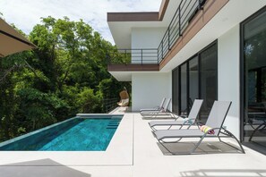 Un área de piscina moderna al aire libre, con una piscina azul cristalina, rodeada de exuberante vegetación y un edificio contemporáneo con grandes ventanales y un balcón.