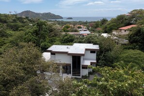 A modern two-story building surrounded by dense greenery, with a view of the sea and hills in the background. - Casa Joanna | Luxury Jungle Retreat in Pacifico Resort | Private Pool (Pacifico Resort Lot 35)