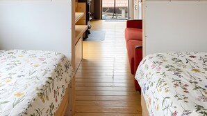 Habitación compacta con dos camas, un sofá rojo, piso de madera y estanterías empotradas con plantas en macetas.