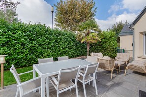 A patio with a table and chairs, surrounded by greenery and a building.