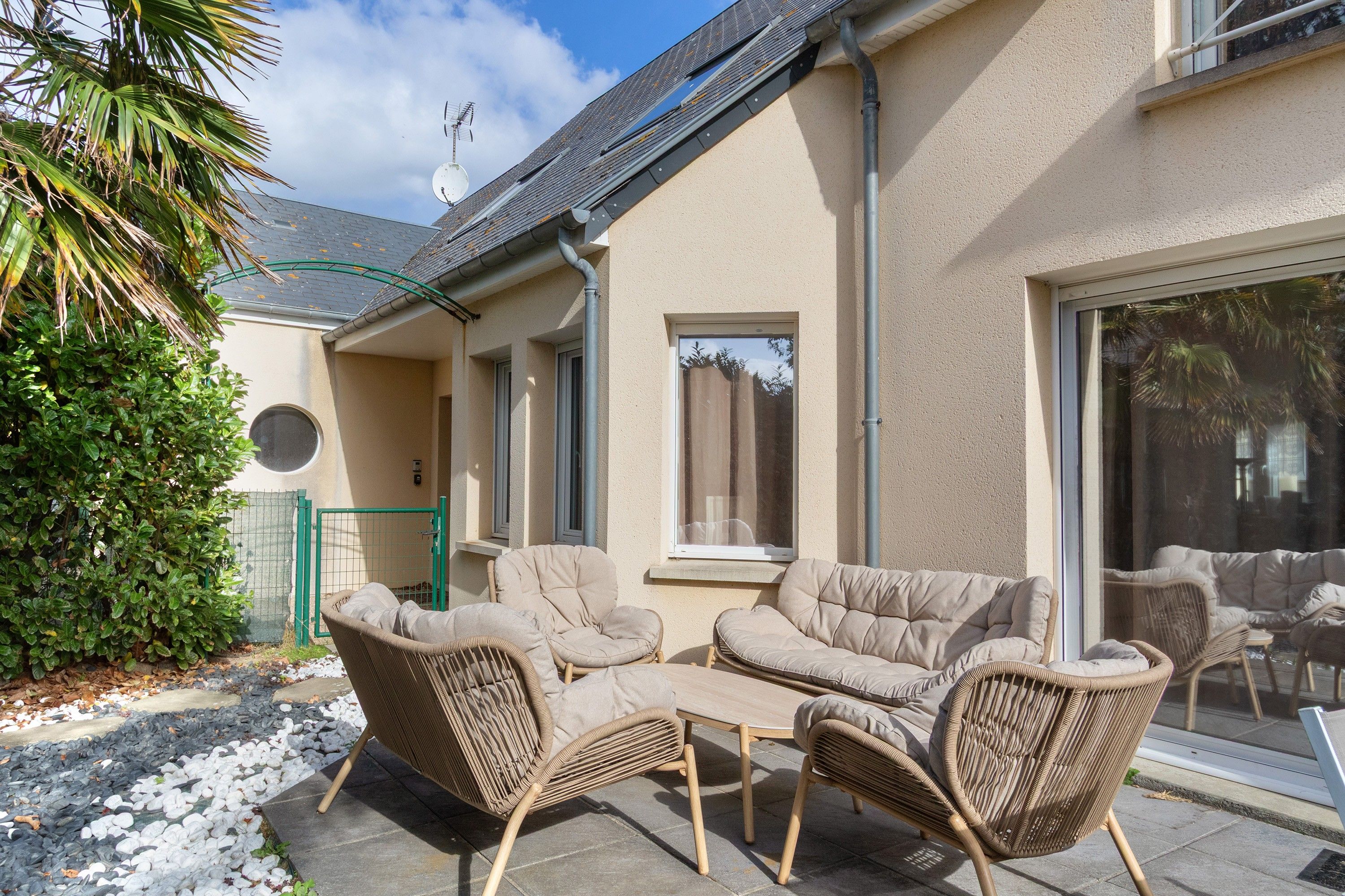 Une terrasse avec des meubles en osier, un portail vert et une maison avec un toit en pente.