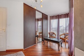 Una habitación con un espejo grande, dos sillas de mimbre, una mesa con cubierta de vidrio, suelo de madera y vistas por la ventana.
