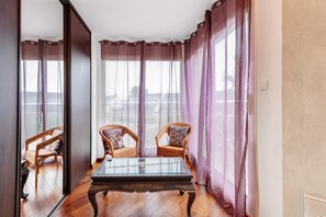 Une pièce avec une table à plateau de verre, deux fauteuils en osier ornés de coussins à motifs, et une vue dégagée sur l’extérieur grâce à de grandes fenêtres et à de fins rideaux.