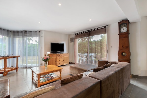 A spacious living room with a large sofa, a wooden coffee table, and a television. There is a dining area with a table and chairs, and a large window with curtains. A wooden clock is visible on the wall.