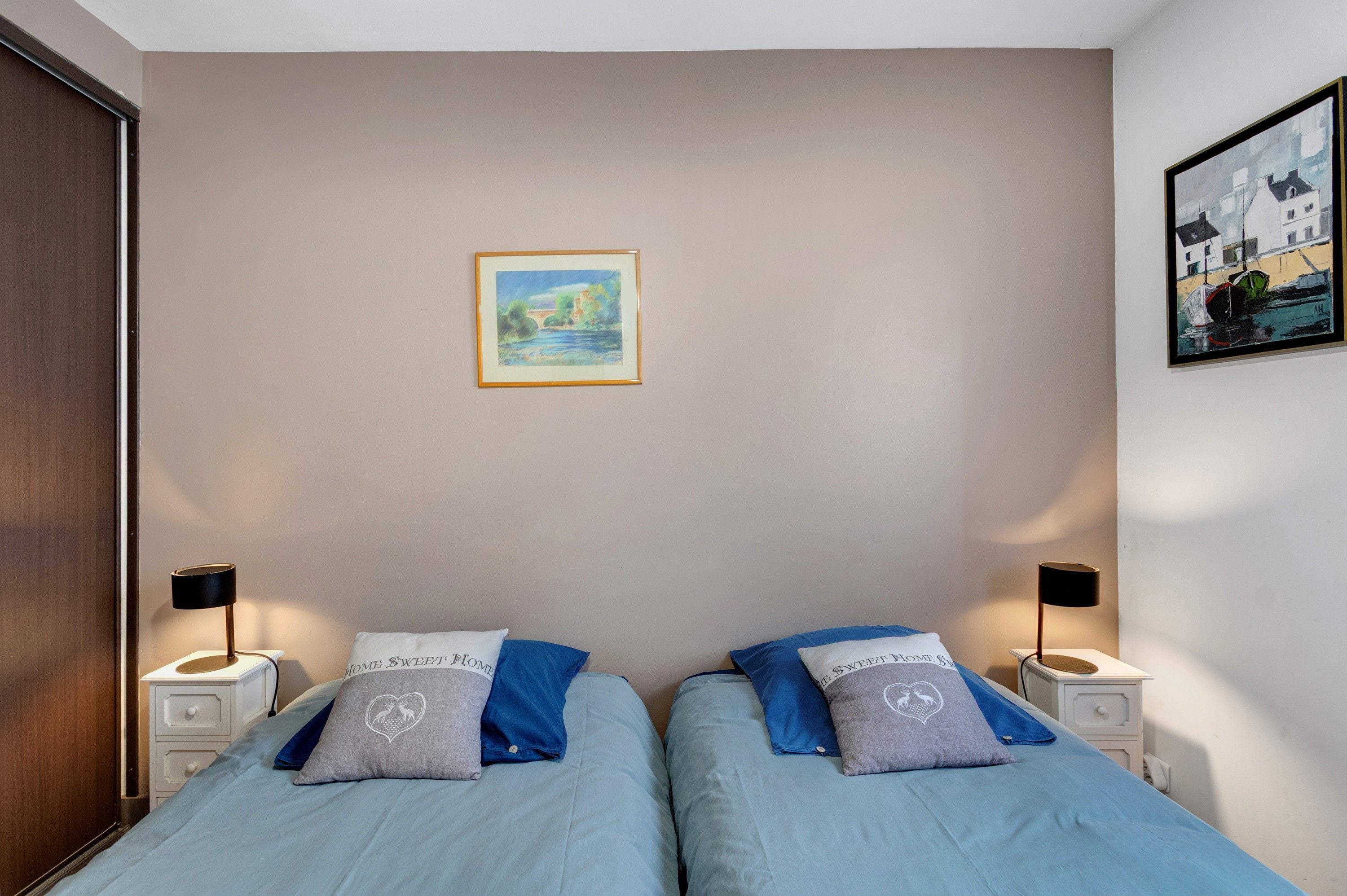 A bedroom with two beds, a nightstand with a lamp, a framed painting on the wall, and a door.