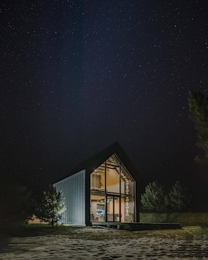 Uma cabana moderna com paredes de vidro e exterior escuro, ao fundo de um céu estrelado.
