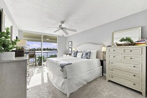 Ein Schlafzimmer mit Bett, Kommode, Deckenventilator und Blick auf ein Gewässer durch eine Schiebetür.