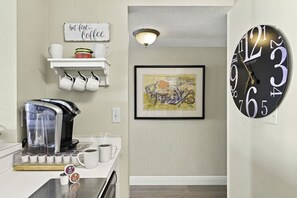 A kitchen with a coffee maker, mugs, and a sign that says 'but first Coffee.' There's a framed painting on the wall and a clock. - Serene Oasis | 2BR Club Bahia Condo | Isla Del Sol (Isla Del Sol)