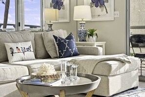 Ein Wohnzimmer mit einer Couch, dekorativen Kissen, einem Couchtisch mit Büchern und einer Vase, und Blick ins Freie.