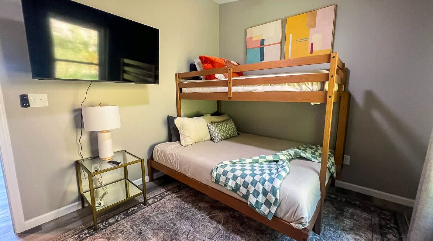 A bunk bed with a television above it, a bedside table with a lamp, and a patterned rug on the floor.