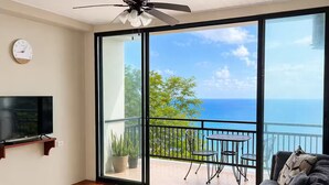 A modern living room with a large grey sofa, a flat-screen TV mounted on the wall, and a balcony with a view of the ocean. - Paramin View- 2 BR Ocean and Mountain Views Retreat (Diego Martin Regional Corporation)