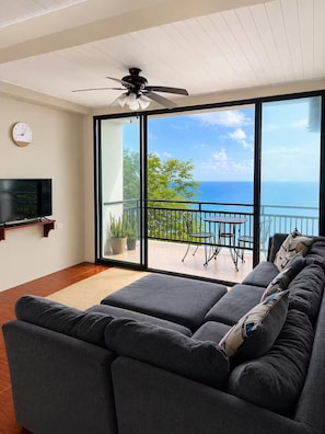 Ein modernes Wohnzimmer mit einer großen, grauen Couch, einem am Wand montierten Flachbildfernseher und einem Balkon mit Meerblick.