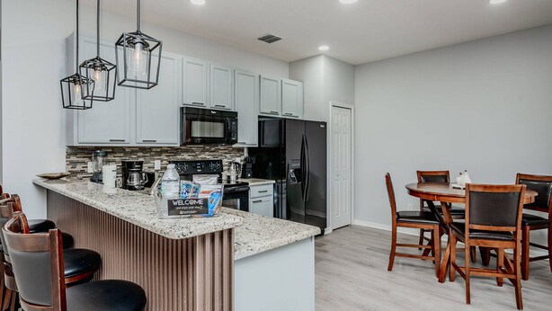 A modern kitchen with white cabinets, a black refrigerator, and a granite countertop. There is a dining area with wooden chairs and a table. - 5BR Veranda Palms Home Near Disney for 14 (Kissimmee)