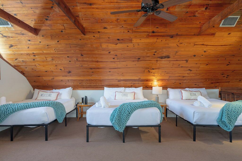 A room with three beds, wooden ceiling beams, a ceiling fan, and a nightstand with a lamp.