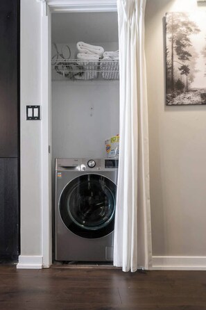 Un cuarto de lavado con una lavadora de carga frontal, un estante con toallas dobladas y un cuadro en la pared.