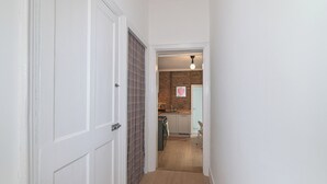 Een smalle gang die leidt naar een keuken met een bakstenen muur, een hangende lamp en een deur met een ruitjes gordijn.