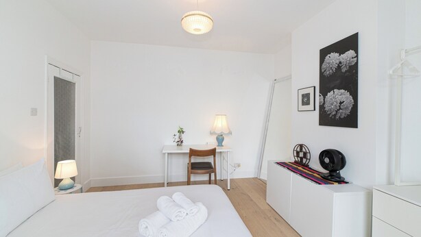 Een hotelkamer met een bed, een bureau, een stoel, een lamp, een fotolijstje en een ventilator.