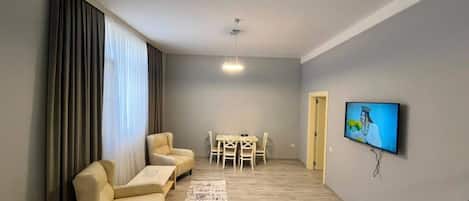 A living room with a sofa, a coffee table, a TV mounted on the wall, and a dining area with chairs and a table.