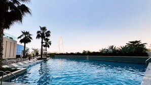 A swimming pool with lounge chairs and palm trees, a Ferris wheel in the background, and modern buildings. - Chic Address JBR Haven w/ Marina View (Dubai)