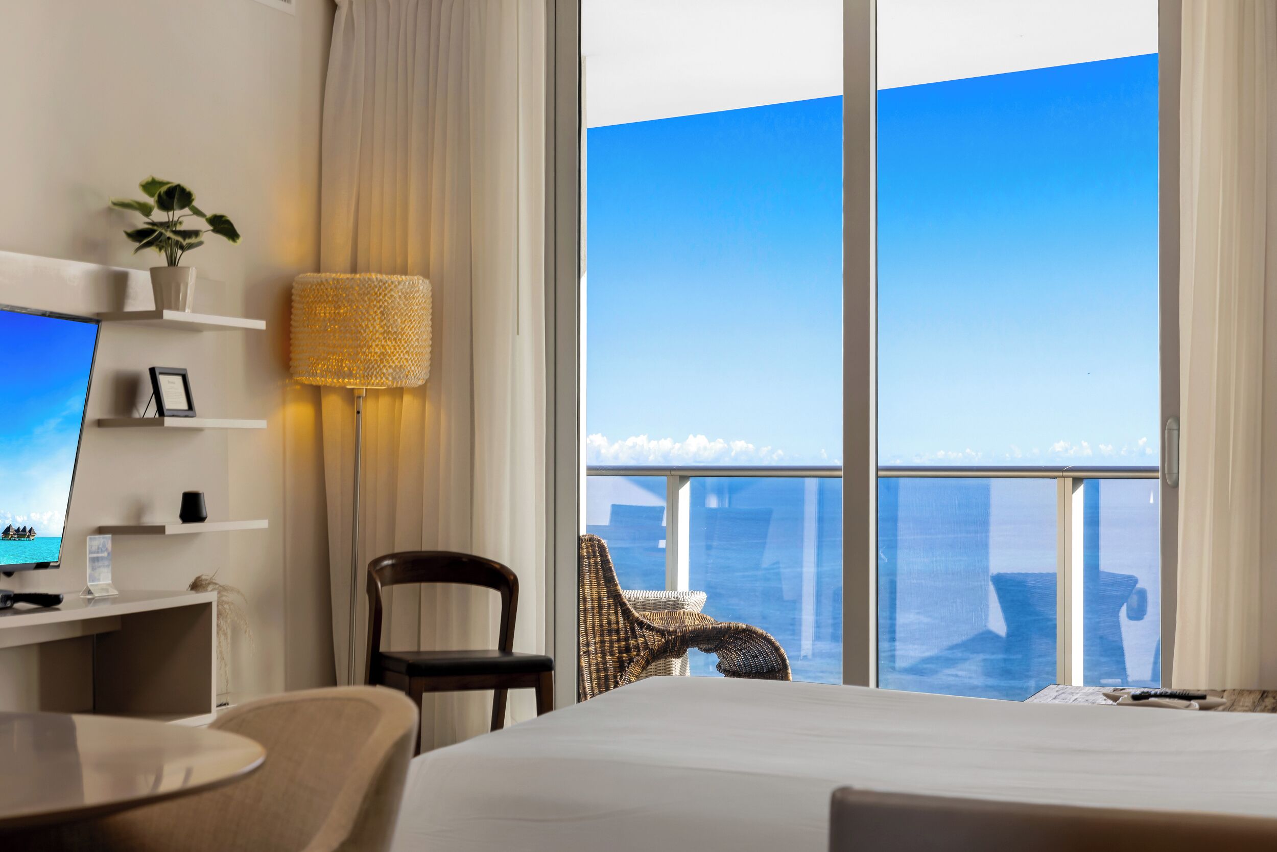A hotel room with a bed, a desk with a computer, a chair, a lamp, and a view of the city through large windows.