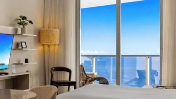 A hotel room with a bed, a desk with a computer, a chair, a lamp, and a view of the city through large windows.