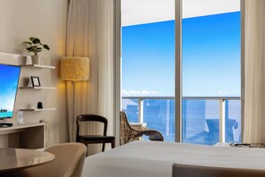 A hotel room with a bed, a desk with a computer, a chair, a lamp, and a view of the city through large windows.
