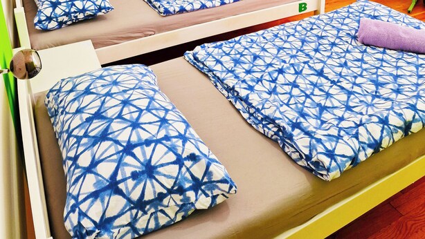 Quarto com duas camas, cada uma com colcha e almofadas estampadas em azul e branco. A parede é verde e há uma pequena mesa com um computador e uma luminária.