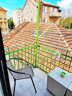 A balcony with a wicker chair and a small table, overlooking a tiled roof and stone buildings.