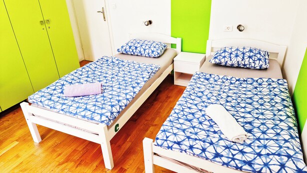 Quarto com duas camas, cada uma com colcha azul estampada e almofadas a combinar, uma pequena mesa de cabeceira branca e um guarda-roupa verde com a inscrição 'Apenas para funcionários'.