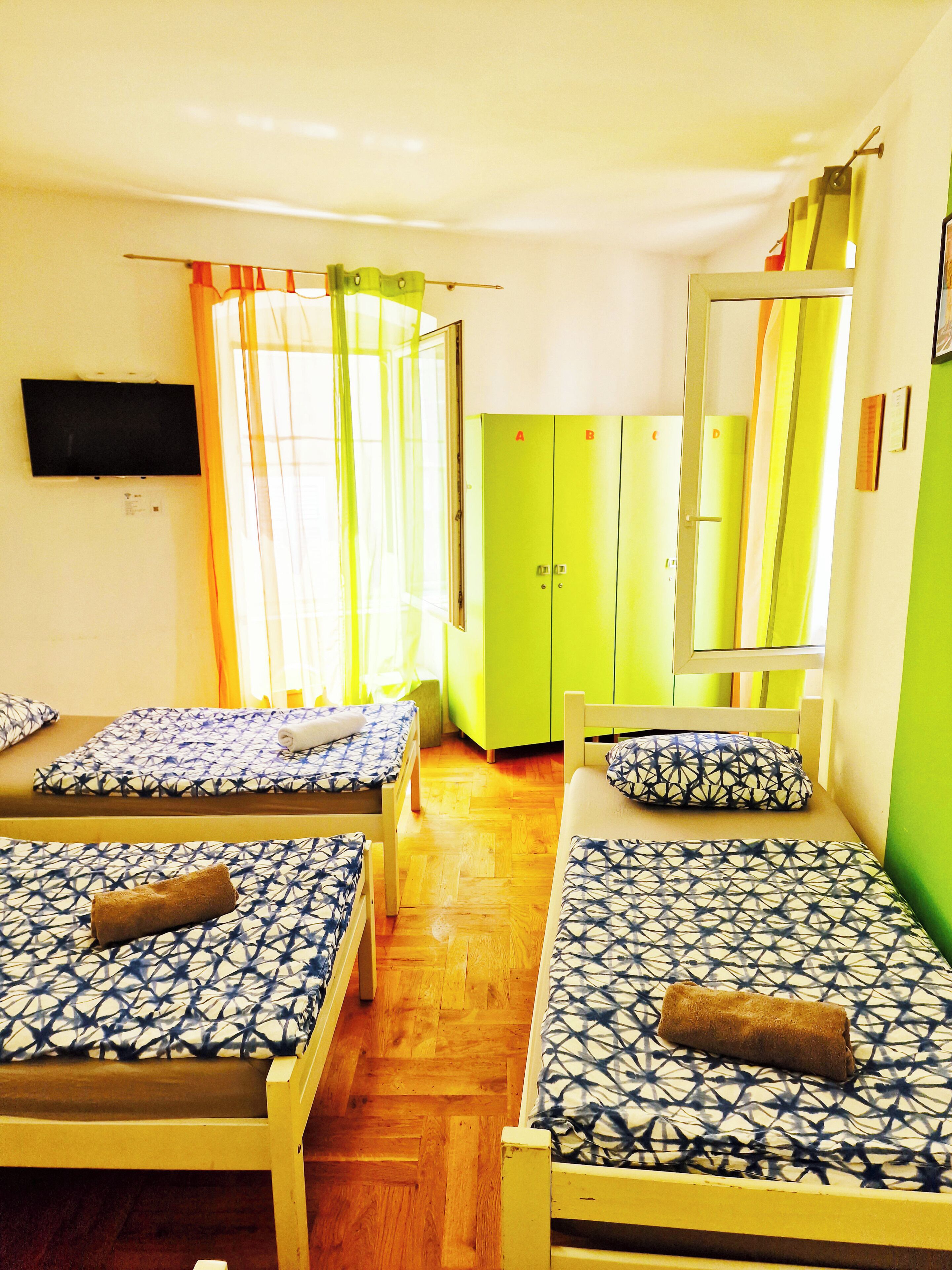 Ein Zimmer mit zwei Betten, einem grünen Kleiderschrank, einem Fernseher und einem Fenster mit Vorhängen.