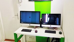 Quarto com uma escrivaninha equipada com dois monitores, teclado e rato. Um quadro colorido está pendurado na parede acima da escrivaninha.