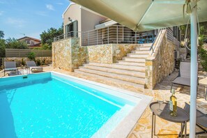 Une piscine avec des marches en pierre menant à un espace couvert, un coin salon extérieur et une vue sur un immeuble d’habitation en arrière-plan.
