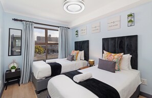 A hotel room with two beds, a large window, and a view of buildings outside. - Cape on Viola | Modern Apartment in Table View (Cape Town)